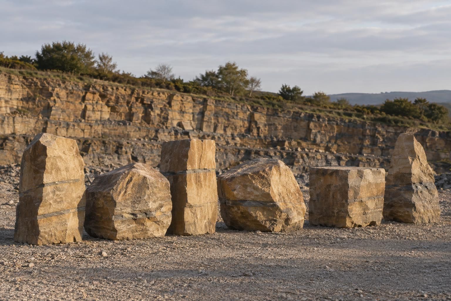 A single line of six large buff sandstone boulders and squarish blocks set out on a gravel quarry floor, each a different shape, each taller than knee height, low evening sun raking across their faces, a stepped quarry face softly out of focus behind.
