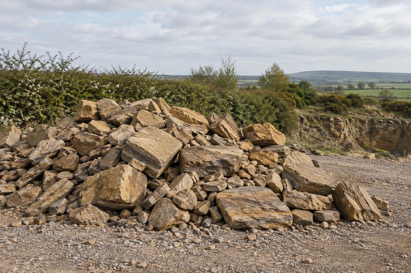 A low, spread heap of mixed rockery-sized sandstone pieces on a gravel floor, varied shapes and weathered faces, a small excavator bucket resting at the edge of frame.